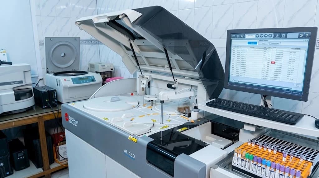 Beckman Coulter AU480 biochemistry analyser with its reagent tray open, sample racks and reporting screen beside it, in the Dhaka Clinic laboratory, Tangail